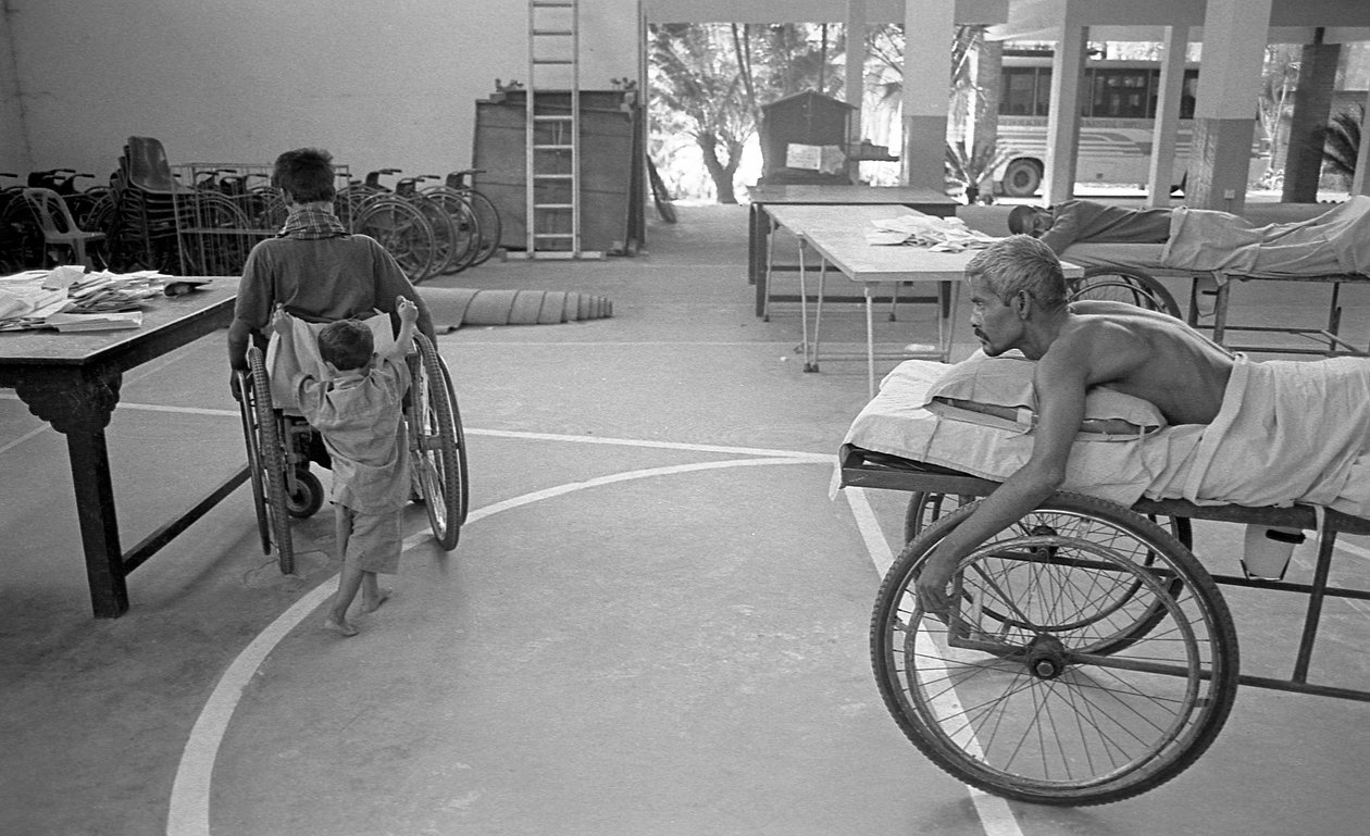 Fyzicky postiženého muže na invalidním vozíku nese jeho syn; další muž ve stejné situaci to sleduje v Centru pro rehabilitaci ochrnutých, CRP v Savar, Dháka, Bangladéš. 2005. od Unbekannt Unbekannt